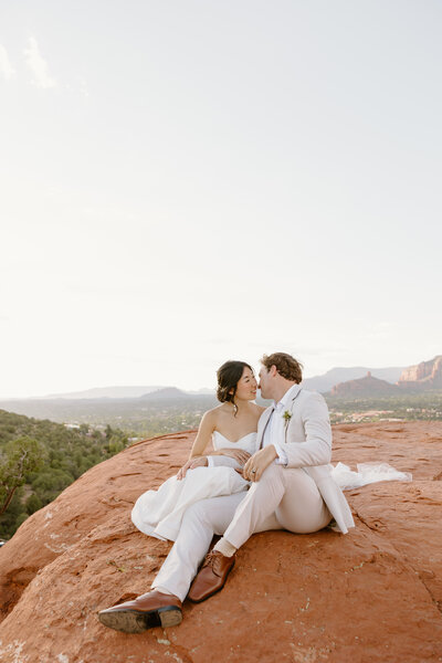 Bride and groom sitting on a rock ledge in their wedding attire leaning in for a kiss.