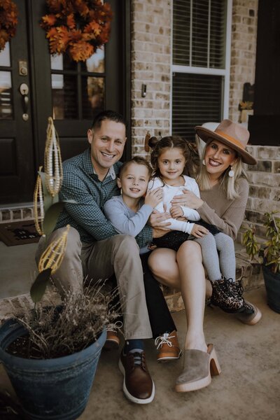 A family of four sits on their front porch, smiling at the camera during a Warner Robins at home family session. The parents sit behind their young son and daughter, while autumn decorations hang on the front door.