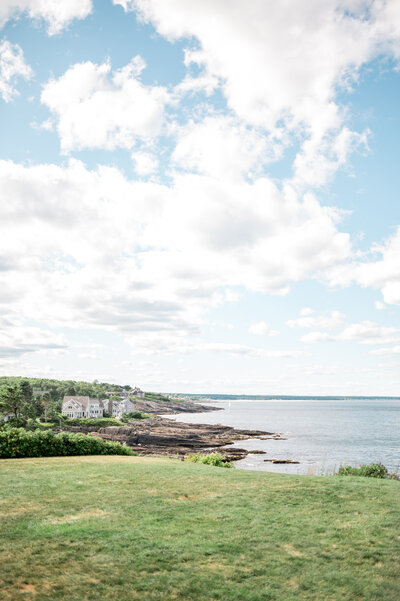 A grassy lawn at Cliff House Maine overlooks a rocky coastline and vast ocean views, with scattered clouds in a soft blue sky.