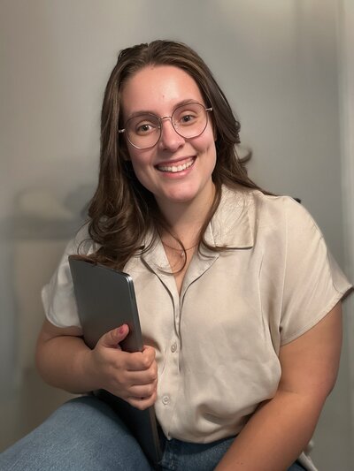 Woman with glasses and brown hair, smiling and holding a laptop, representing approachable virtual assistant and web design services.