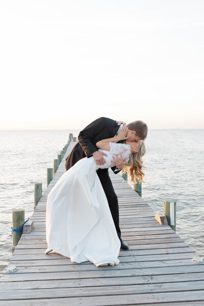 Photo of a groom dipping his bride for a kiss on a pier over Mobile Bay at sunset.