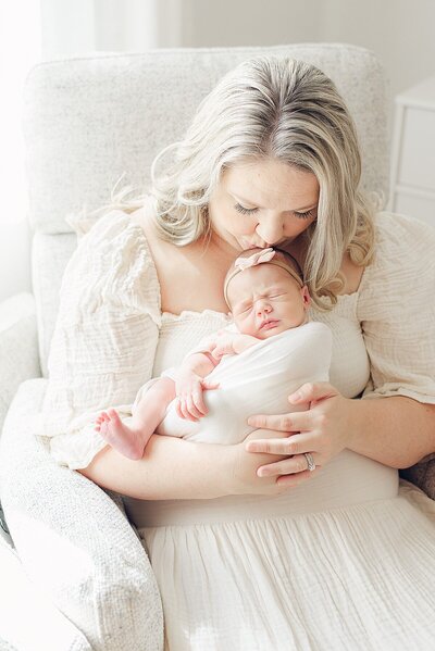 Phoenix mom holding newborn baby facing her while standing in front of window for in-home session. At her feet is an English Bulldog looking at camera.