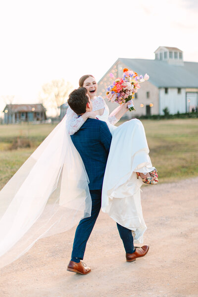 Groom carrying bride while she holds her bouquet and her veil trails on the ground
