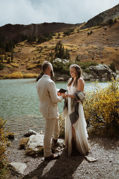 bride and groom exchanging vows at an alpine lake. they are smiling as the sun peaks out from behind the mountains. The bride has mud on the bottom of her dress and is holding her vow book. the groom is wearing hiking boots and a tan jacket.