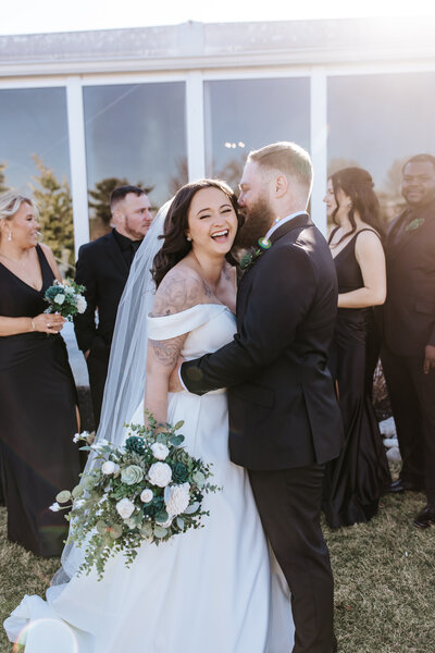 Bride laughing as groom kisses her cheek during golden hour portraits with their wedding party in black attire, captured in candid, joyful style.