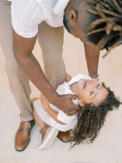 Black dad in white shirt and khakis reaches down and tickles long and curly-haired son attached to his leg photographed by photographer Little Rock Bailey Feeler Photography