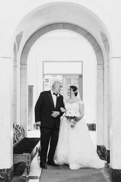 A bride walks arm in arm with her father beneath the stone arches of Saints Peter and Paul Jesuit Church in Detroit, captured in a timeless black and white moment before the ceremony.