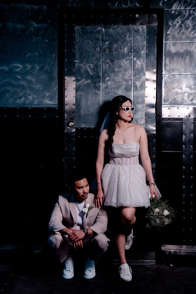 Bride in short white dress holding bouquet and groom in light suit standing against a dark textured wall, modern wedding portrait photographed in Lincoln, Nebraska by Legacy in Light Photography.
