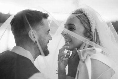 A women smiles and laughs underneath a veil as her fiance says something to her.