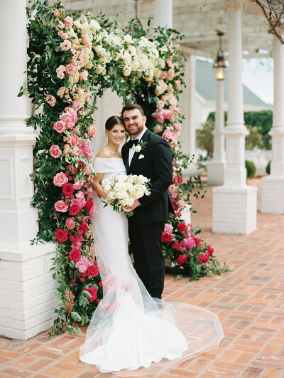 Bride and groom posing in front of a large wall of greenery and pink flowers