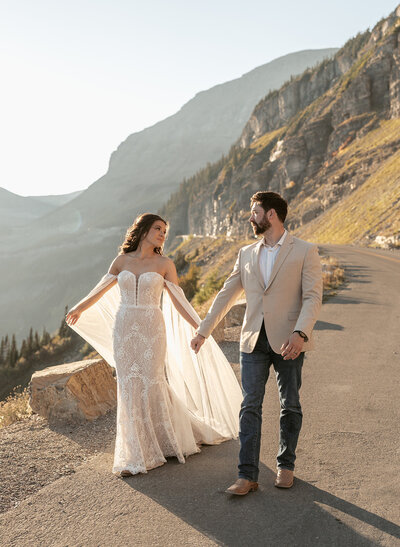 A bride and groom walk hand in hand along a winding mountain road at sunrise in Glacier National Park, the bride’s lace gown catching the golden light, photographed by Sydney Breann Photography.