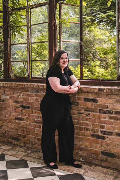 Florist in black pantsuit leaning against wall in Sekrit Theater greenhouse during branding session in Austin, TX.