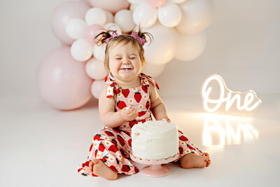 Baby girl with pigtails and dressed in a strawberry jumper eating cake.