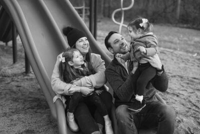 A black and white photo of a joyful family sitting at the bottom of a slide in a playground. A woman and a little girl with a bow in her hair sit on one side. A man holds a small child with a bow, looking at her. They all share a happy moment together.