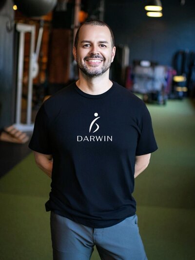 Darwin Ruiz smiling in a black T-shirt featuring the centered “Darwin” logo and motion-inspired icon, standing confidently in a modern gym setting.