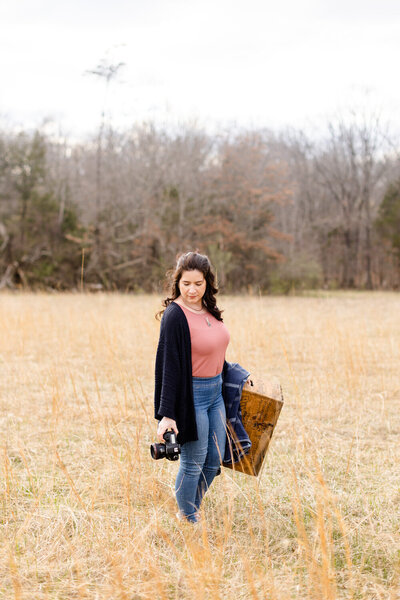 woman holding a camera and a box while walking through a field