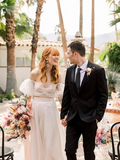 Kohnur couple in a Palm Springs courtyard. Bride in a two-piece off-shoulder dress with red fringe hair and colorful bouquet.