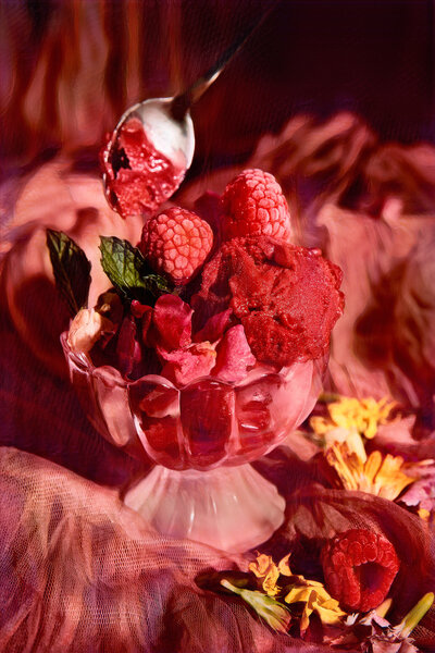 A rich and vibrant image of raspberry sorbet served in a glass dish, garnished with fresh raspberries and mint leaves, surrounded by decorative flowers. Photographed by Mica McCook, this scene is a feast for the eyes, showcasing the deep red hues and luscious textures of the dessert.