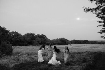 Black and white landscape image of bride and her bridesmaids holding hands and spinning around in a grass field