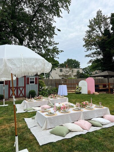 Chic backyard celebration with low picnic tables, pastel cushions, floral centerpieces, and a white fringe umbrella, styled by Yours Truly, a wedding planner and designer in New Jersey.