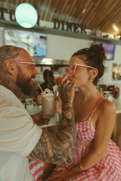 Tattooed couple wearing heart-shaped sunglasses sharing a milkshake at a vintage-style diner, laughing and flirting in a playful retro-themed photoshoot.