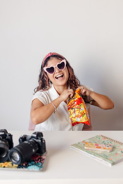 latina woman photographer wearing pink heart sunglasses laughs as she opens a bag of flaming hot cheetos