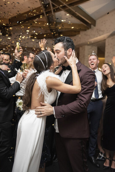 Bride and groom share a kiss on the dance floor surrounded by cheering guests. The bride is wearing a white, backless dress with feather details, and the groom is in a brown tuxedo. Gold confetti is falling from above.