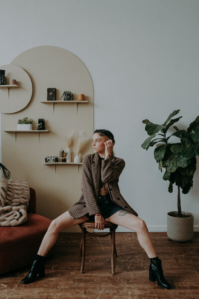 Photographer sits confidently on a wooden chair wearing a vintage blazer and boots, in a warm and styled studio space with plants and retro decor.