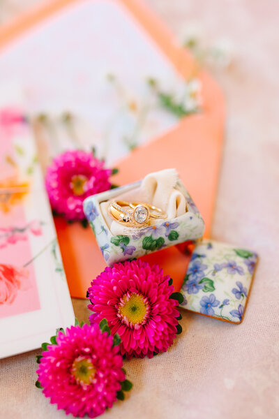 Vibrant photo of wedding rings in a floral ring box next to pink flowers and bright stationery