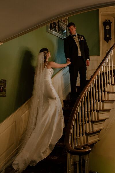 A bride in a white dress and long veil holds hands with a groom in a black suit on a vintage staircase, conveying a romantic, timeless moment.