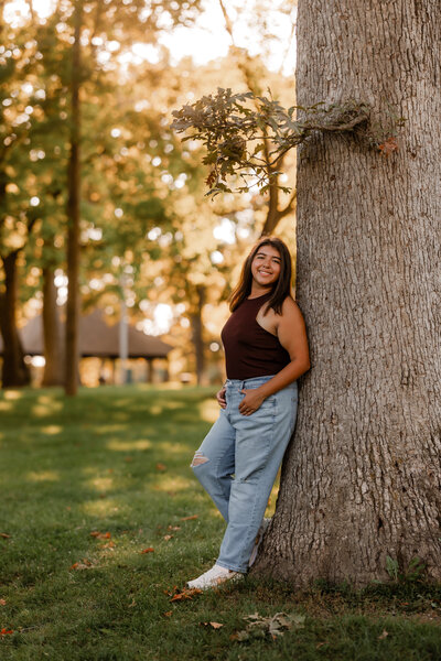 A young woman poses against a tree during sunset for senior photos.
