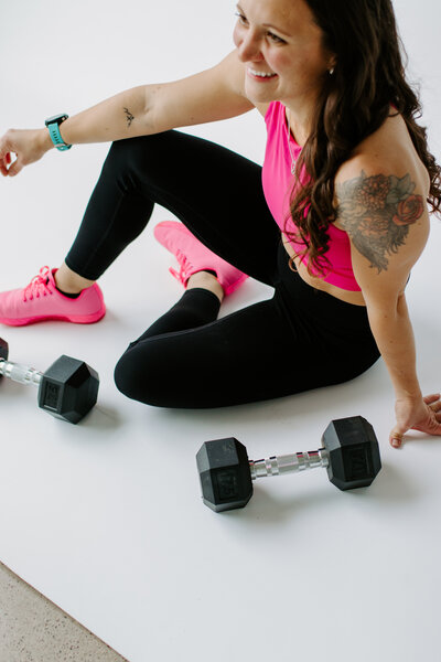Woman with dark hair sitting on ground in black leggings, pink sports bra and pink shoes with weights