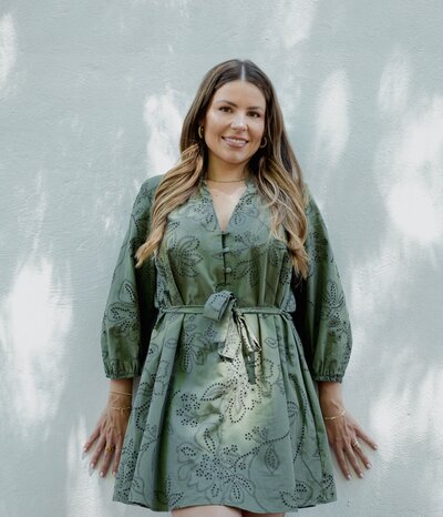 Natalie Bernacchi standing against a light-colored textured wall, smiling softly, wearing a belted olive green eyelet dress with gold jewelry and long wavy hair offering real-time mindset, business, and energetic support through private messaging and voice notes.