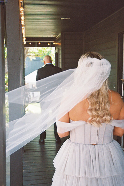 A bride's vail blows in the wind as she approaches the groom, representing the film wedding photography style of Among the Oaks. 