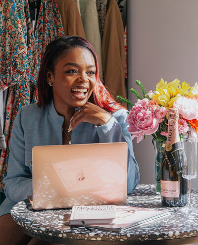 Tahirah sitting at her laptop smiling with a bouquet and a bottle of Moet & Chandon next to her