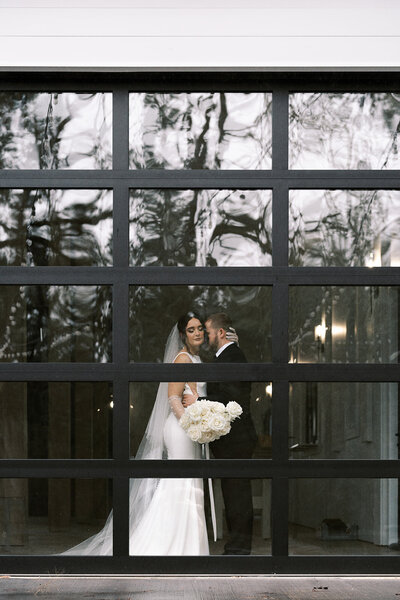 A bride and groom embrace through a window pane. 