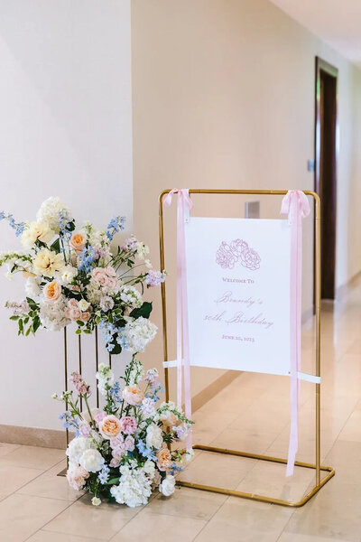 delicate line art floral welcome sign tied with pink ribbon on each side displayed with a golden stand next to pastel florals