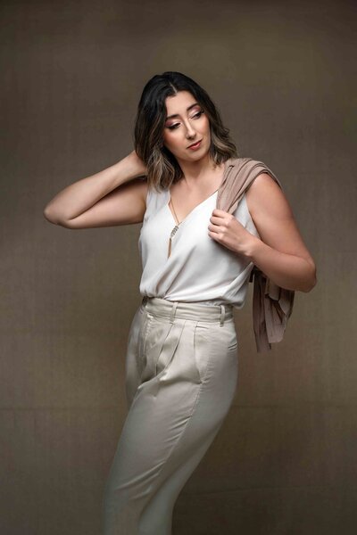 Tacoma woman poses for artistic portrait during headshot session in studio vogue aesthetic cardigan over shoulder cream top khaki slacks touching hair