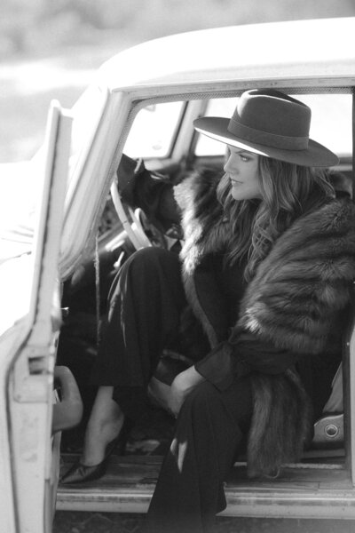 Jess Kuipers, Santa Barbara wedding planner, wears fur stole and hat while sitting in drivers seat of vintage car