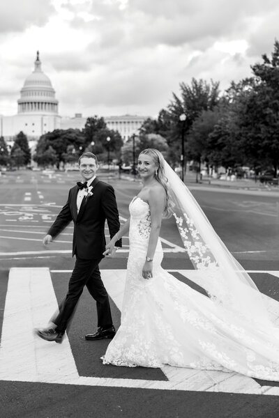 A couple in wedding attire holding hands and smiling while walking on a crosswalk in front of the United States Capitol building. The bride wears a long, lace veil and a gown, and the groom is in a dark suit with a bow tie. Trees and cloudy skies are in the background.