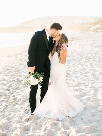 A bride and groom share a romantic kiss on the sandy shores of Laguna Beach at sunset, celebrating their Ritz-Carlton Laguna Niguel wedding