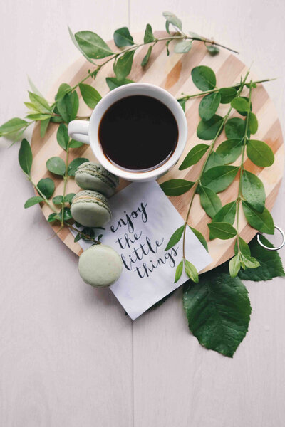 Cup of coffee with macarons, leaves and a note that says "enjoy the little things"