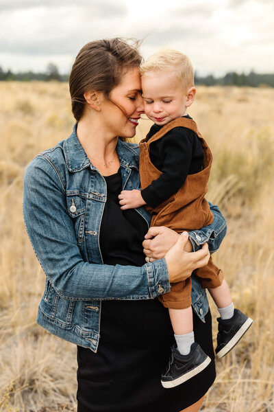 brunette mother in jean jacket nuzzling blonde little boy in brown overalls