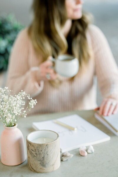 A woman holding a coffee mug at a desk with a notebook, candle, vase of baby's breath, and healing crystals.