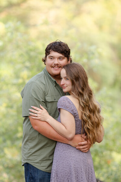Engaged couple with the man's hands around her waist, both looking off camera against a creamy green tree background.
