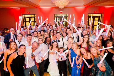 Wedding party crowd celebrating with raised hands and glow sticks on a vibrant dance floor.