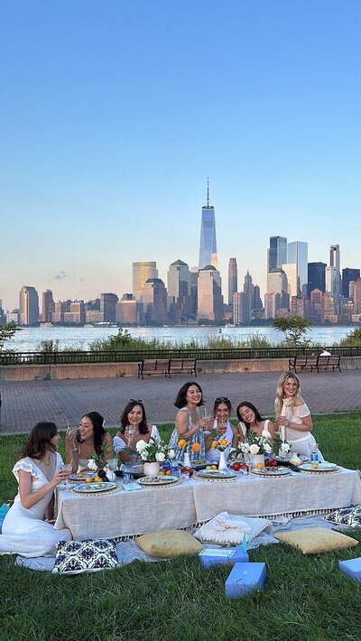 Group of women enjoying a styled outdoor dinner picnic with floral décor and views of the New York City skyline, designed by Yours Truly, a wedding planner and designer in New Jersey.
