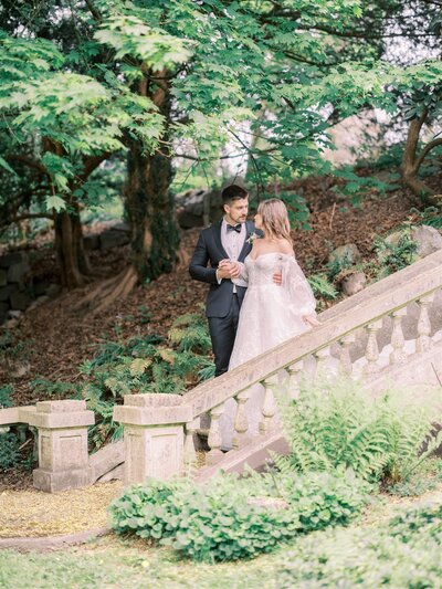 A bride and groom in wedding attire stand on a stone staircase surrounded by lush greenery. The groom holds the bride's hand as they gaze at each other lovingly, their moment perfectly orchestrated by a top-tier wedding planner in Canada.