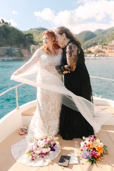 Lelia Angelique and her wife Sarah standing on a boat during their wedding day, with Sarah kissing Lelia's forehead and a stunning ocean and coastal backdrop behind them.