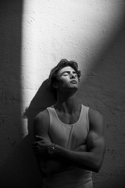 Striking black-and-white portrait of a teen guy in a white tank top, standing in dramatic lighting with shadows on a textured wall—strong editorial feel.